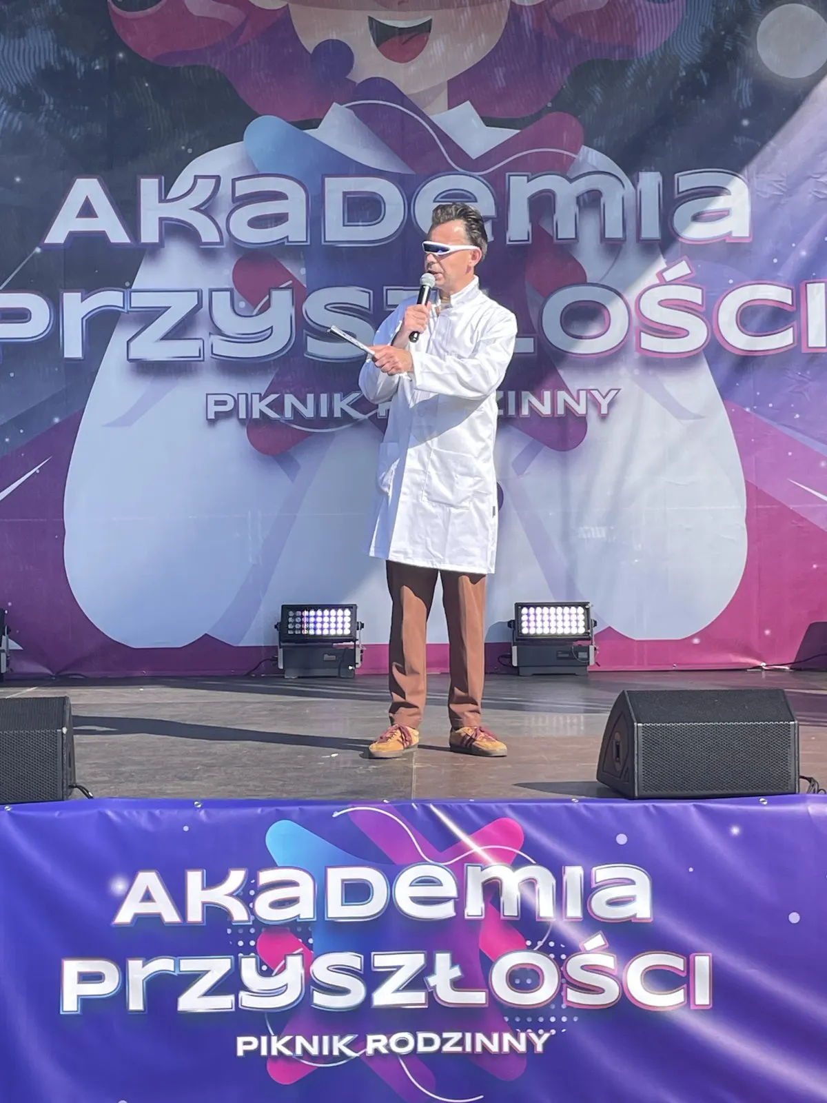 Konferansjer w białym fartuchu naukowca z mikrofonem na scenie pikniku Akademia Przyszłości Sieci Badawczej Łukasiewicz