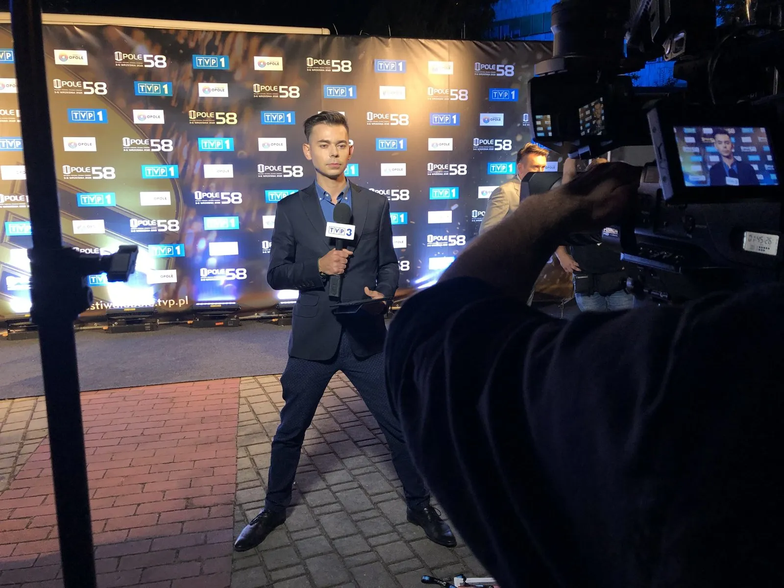 Szymon Alaba z mikrofonem przed ścianką 58. Festiwalu w Opolu podczas wejścia na żywo, obok kamera telewizyjna