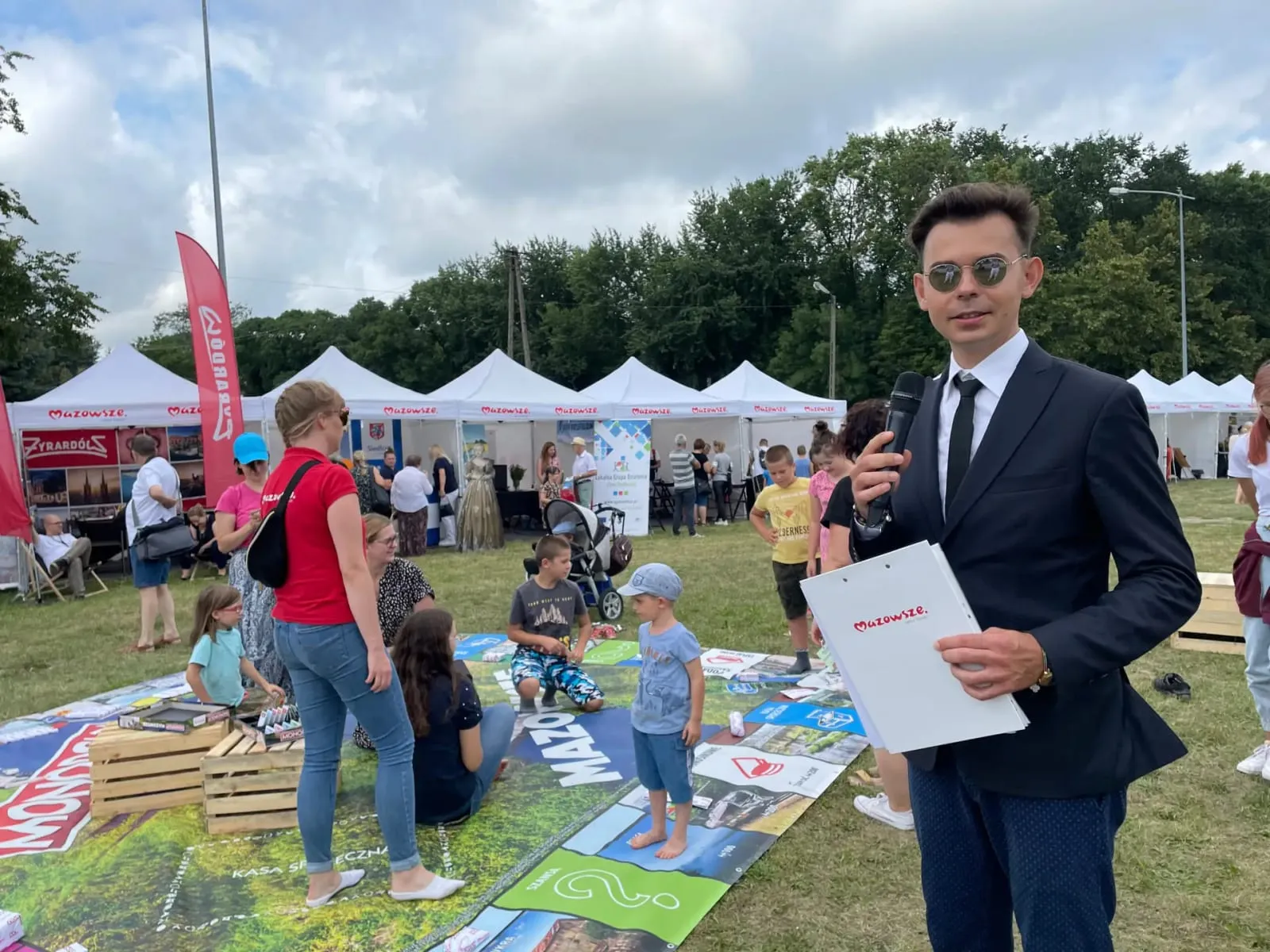 Prowadzący z mikrofonem obok wielkoformatowej planszy Monopoly Mazowsze na Mazopikniku
