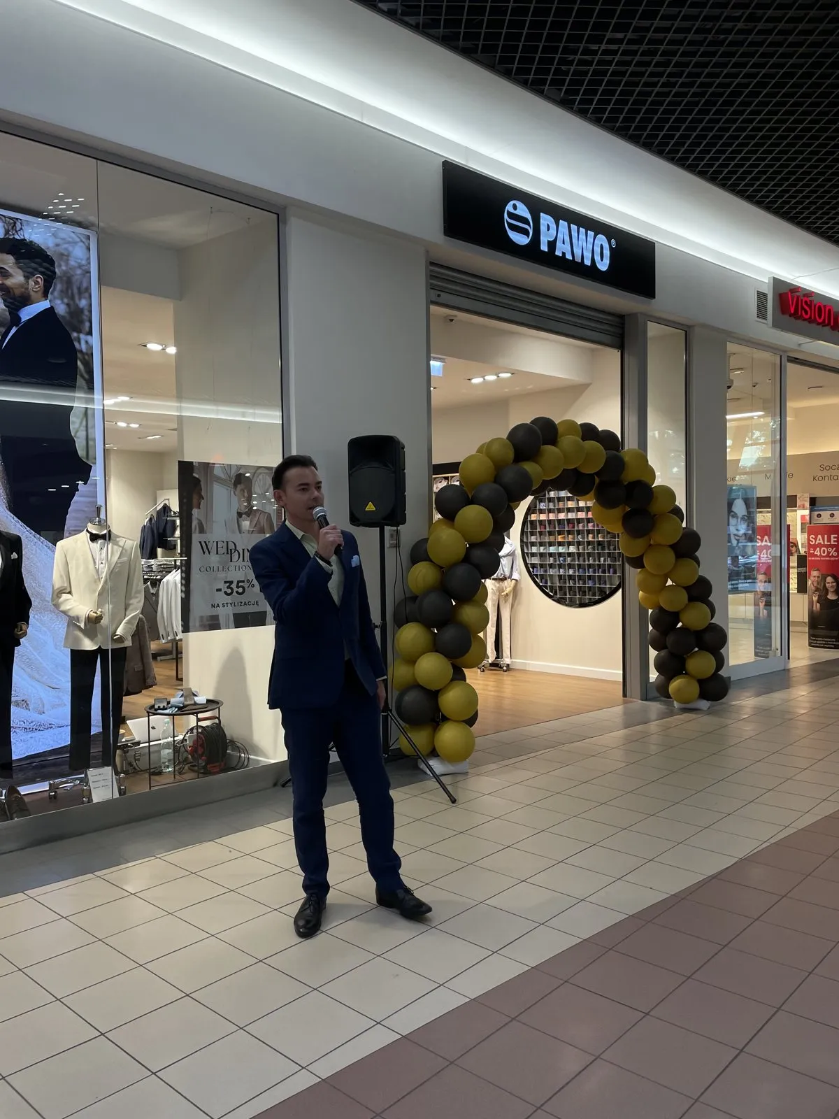 Konferansjer w garniturze z mikrofonem przed wejściem do salonu PAWO z balonową bramą