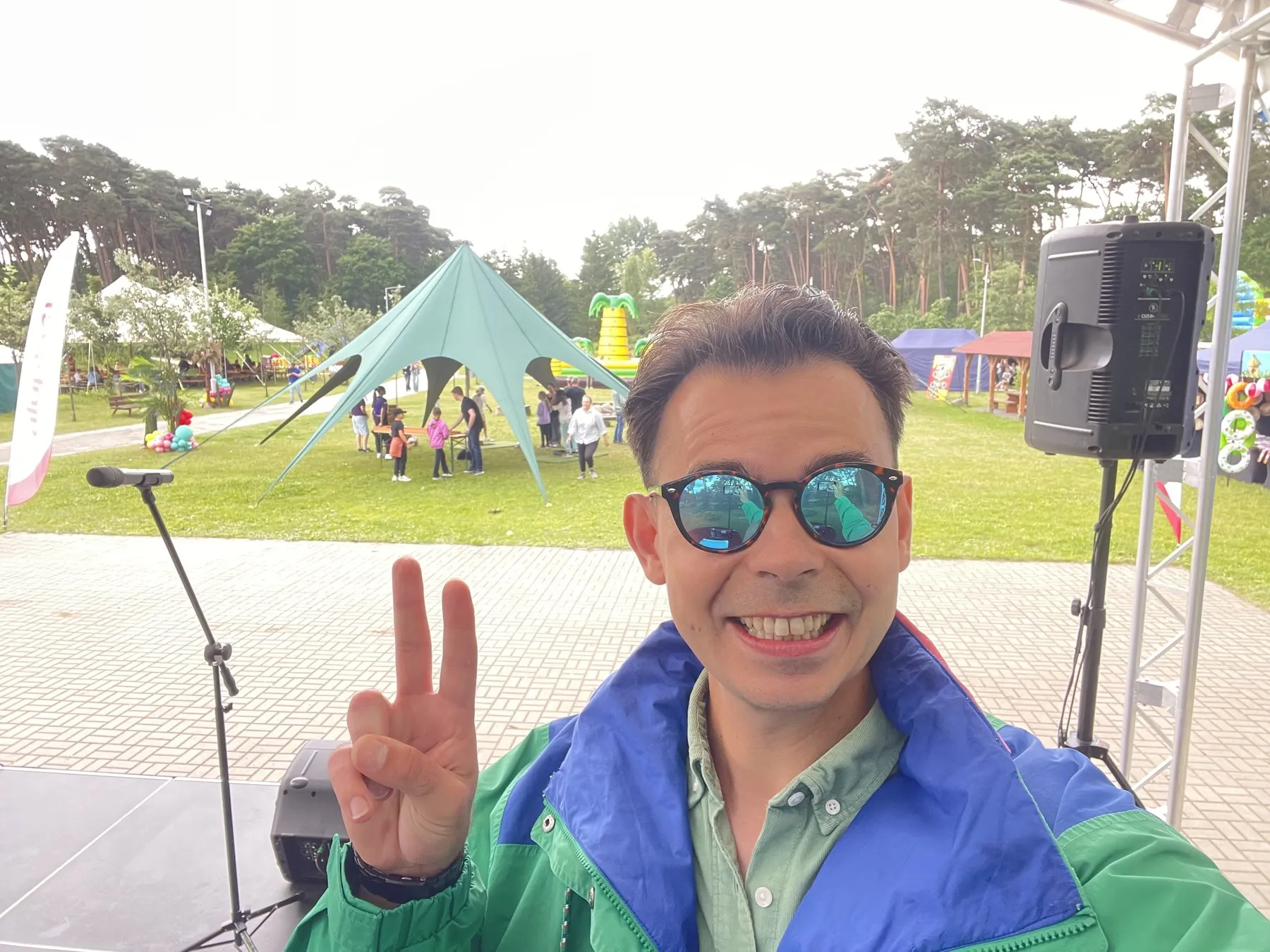 Konferansjer Szymon Alaba na scenie pikniku aluplast w Ogrodzie Dendrologicznym w Poznaniu, w tle atrakcje dla rodzin