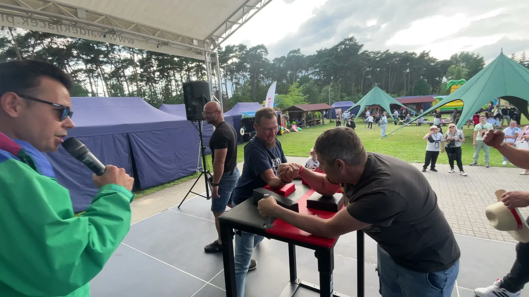 Konferansjer z mikrofonem komentuje turniej siłowania na rękę na scenie pikniku aluplast